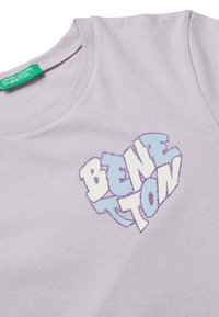 Camiseta de algodón de color púrpura claro con un logo en forma de corazón "BENETTON" en blanco con acentos de purpurina azul y púrpura. La etiqueta dice "United Colors of Benetton."