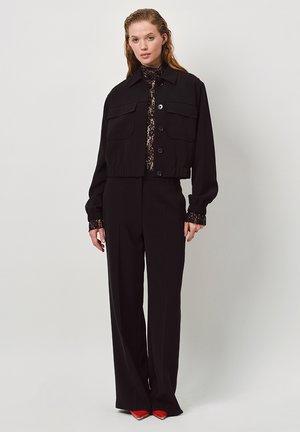 Zwarte cropped jas met twee voorzakken, gecombineerd met een doorschijnend zwart kanten top, en wijde zwarte broek. Rode pointed shoes accentueren de outfit.