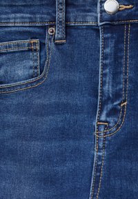 Gros plan d'un jean en denim bleu montrant la poche avant, la boucle de ceinture, des coutures piquées et un bouton métallique argenté à la ceinture.