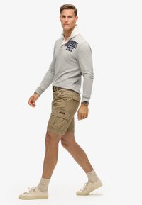 Polo gris à manches longues avec col blanc et lettrage marine, associé à un short cargo beige et des baskets légères. Texture douce, coupe décontractée.