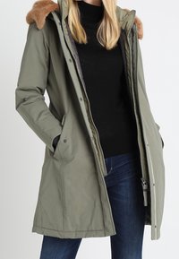 Olivově zelená parka s kapucí lemovanou umělým kožíškem, zapínáním na zip a bočními kapsami. Má mírně delší siluetu s hladkou texturou.