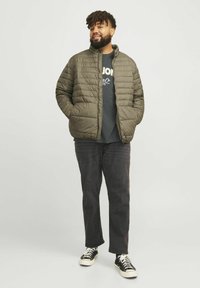 Olijfgroene pufferjack met horizontale stiksels, een opstaande kraag, gecombineerd met een donker t-shirt en zwarte spijkerbroek. Sneakers met witte zolen.