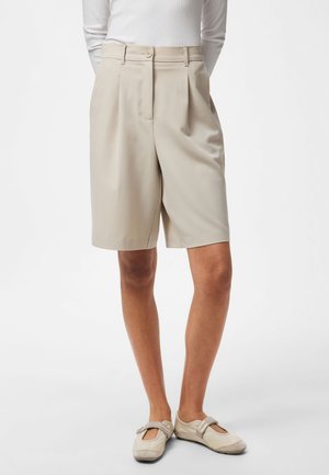 Short sur mesure beige coupé au niveau du genou, porté avec un top blanc à manches longues et des chaussures plates beige style Mary Jane.