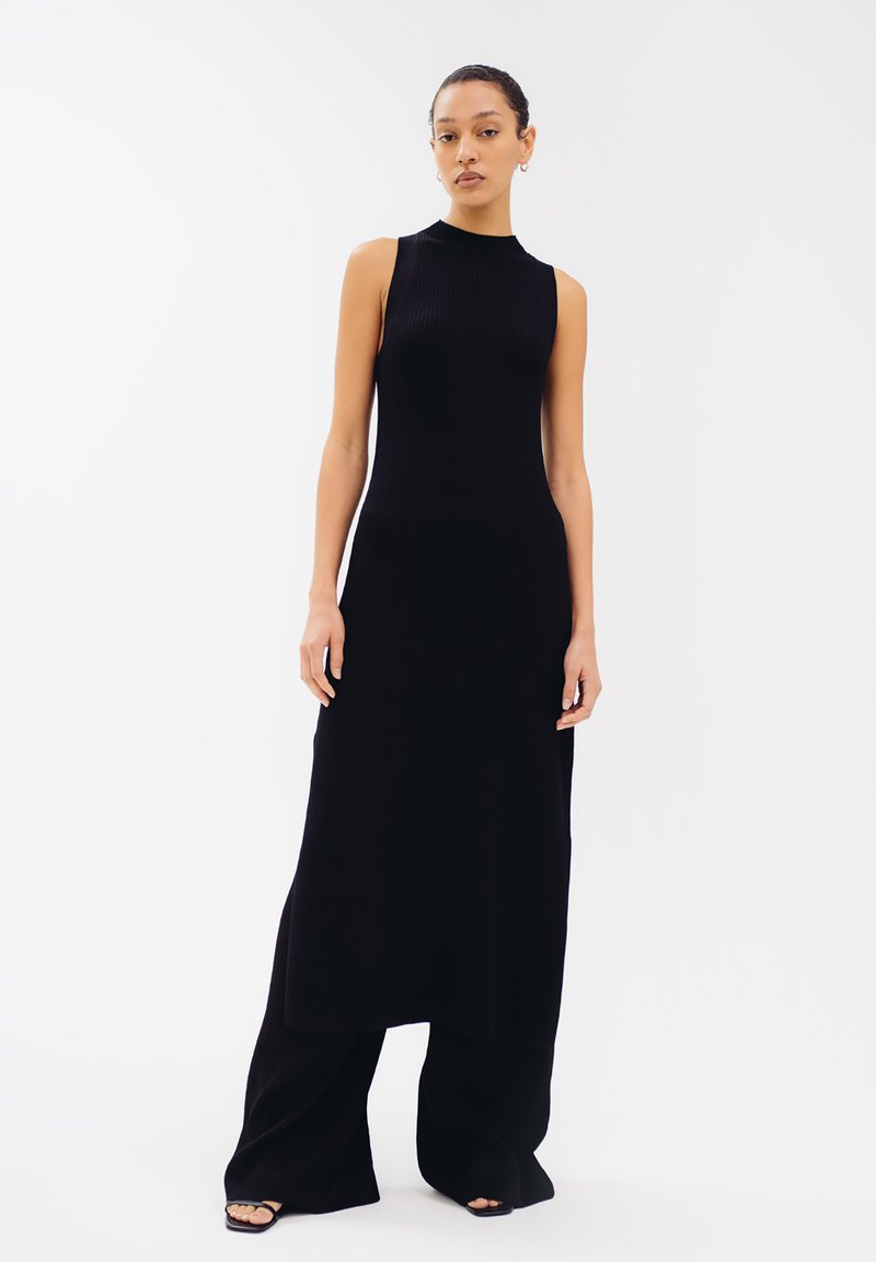 Schwarzes, geripptes, ärmelloses Kleid mit hohem Ausschnitt und gerader Silhouette, kombiniert mit weiten schwarzen Hosen. Glatte Textur und bodenlang.