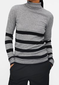 Pull à col roulé gris avec des rayures noires horizontales, doté d'une texture en tricot doux et de poignets côtelés. Assorti à un pantalon noir.