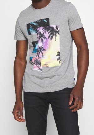 Mann trägt graues kurzärmeliges T-Shirt mit bunten, mehrlagigen Palmenmotiven und schwarze Hose, steht vor schlichtem Hintergrund.