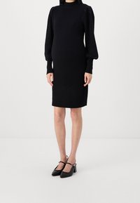 Robe tricotée noire à côtes, à coupe slim, avec un col montant et des manches ballon. Associée à des escarpins noirs à talon bas.