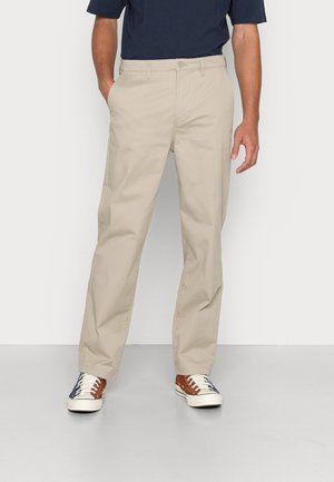 Chinos in cotone beige con taglio dritto. Presentano una chiusura con bottoni, tasche laterali e una texture liscia. Abbinati a sneakers multicolori.