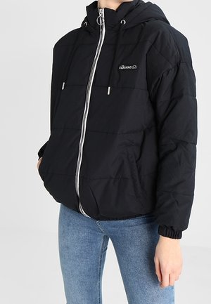 Zwart pufferjack met capuchon, zilveren ritssluiting, ellesse-logo op de borst, gedragen met blauwe spijkerbroek, handen in de zakken, staand tegen een effen achtergrond.