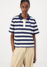Polo a righe navy e crema con colletto a bottoni e un piccolo logo sul lato sinistro del petto. Maniche corte e vestibilità rilassata.