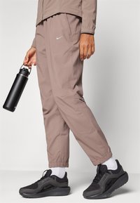 Lichtbruine Nike-broek met elastische boorden, zakdetails, gecombineerd met zwarte Nike-sneakers en een zwarte waterfles.