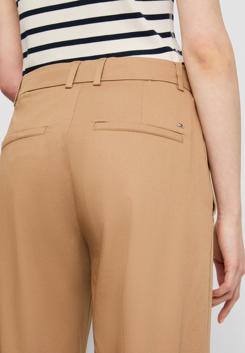 Pantaloni bej cu o textură netedă, având brâie pentru curea și două buzunare la spate. Există un mic eticheta cu logo multicolor pe talie.