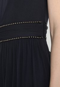 Dunkelblaues Kleid mit plissiertem Design, tailliertem Schnitt und einer dekorativen Reihe von metallischen Perlen entlang der Naht. Weicher Stoff.