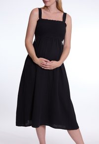 Schwarzes Umstandskleid mit gerafftem Oberteil und breiten Trägern, das eine fließende A-Linie hat und aus strukturiertem Stoff besteht, der bis zur Mitte der Waden reicht.
