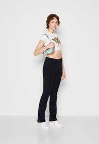 Hvit kortermet t-skjorte med sommerfugl- og dyremotiv, kombinert med mørkeblå jeans og hvite joggesko. Modellen holder en liten iriserende veske.