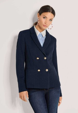 Vrouw draagt een getextureerde marineblauwe dubbelrijige blazer met gouden knopen over een blauw-wit gestreept overhemd met kraag en donkerblauwe jeans.