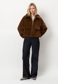 Bruine faux bontjas met een rits aan de voorkant, brede kraag en twee voorzakken, gecombineerd met donkerdenim flared jeans en gestippelde schoenen.