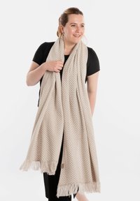Beige geweven sjaal met een gevarieerd patroon, franjes aan de randen, van zacht materiaal. Gedrapeerd om een persoon in een zwarte top.