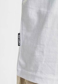 Camiseta gris claro de algodón, con una pequeña etiqueta negra que lleva el texto "CAYLER" en el lado izquierdo. Textura suave y corte informal.