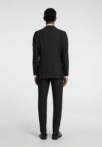 Traje gris oscuro, diseño ajustado, tejido texturizado, con una abertura trasera y cierre de dos botones. Puños de camisa blanca arremangados y zapatos negros.