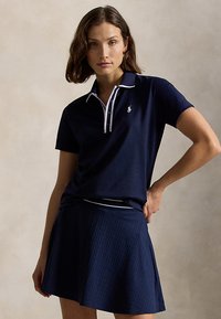 Kvinne iført marineblå kortermet poloshirt med hvit kant og matchende teksturert marineblå plissett skjørt, stående med hånden på hoften.