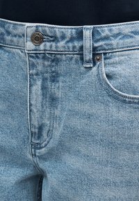 Šviesiai mėlynos spalvos džinsai iš džinsinio audinio, susegami sagomis, penkių kišenių dizainas ir subtilus išblukimas. Audinys turi tekstūruotą paviršių su matoma siūle.