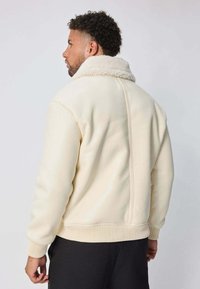 Veste bomber de couleur crème avec un col texturé et duveteux. Présente une base et des poignets côtelés, avec une finition en tissu lisse et un design minimaliste.