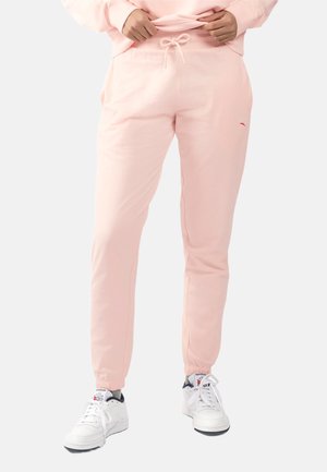 Handen die lichtroze joggerbroeken met trekkoord aanpassen, gedragen met witte Reebok-sneakers en grijze sokken op een effen achtergrond.