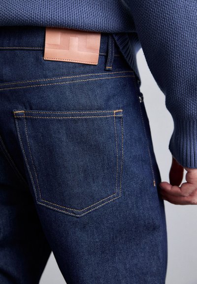 Jeans de mezclilla azul oscuro con costuras naranjas, que cuentan con un bolsillo trasero y una etiqueta de cuero marrón claro en la cinturilla. Textura suave.