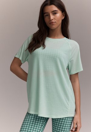 Femme aux longs cheveux bruns portant un t-shirt ample vert clair et un pantalon vert à carreaux, debout devant un fond neutre.