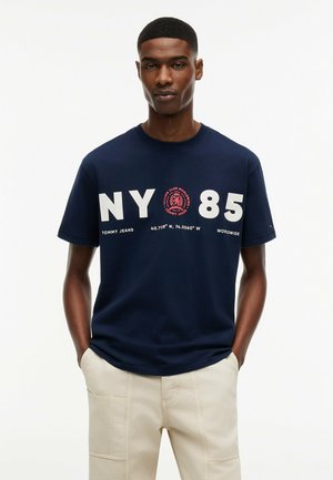 Mand iført navy blå NY 85 grafisk t-shirt og lyse beige bukser, stående med hænderne i lommerne mod en ensfarvet hvid baggrund.
