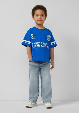 Małe dziecko o kręconych włosach, ubrane w niebieską koszulkę piłkarską "Future Stars Play!", jasnoniebieskie dżinsy i białe tenisówki, stojące pewnie.