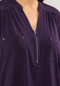 Lila Tunika mit Reißverschluss vorne, zwei Brusttaschen mit metallischen Akzenten und weichem, glattem Material mit gerafften Details an den Schultern.