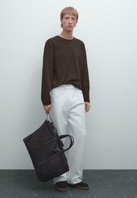 Lange mouw bruine top, witte wijde broek, houdt een grote donkere tas vast. De tas heeft dubbele handvatten en een ritsluiting.