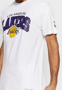 Camiseta blanca usada por una persona, con el texto "Los Angeles Lakers" y un logo de baloncesto amarillo, además del logo de la NBA en la manga.