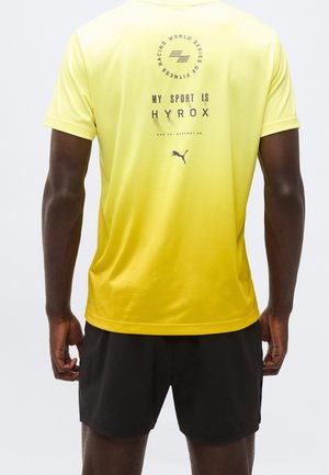 Homme portant un t-shirt de sport jaune avec le texte "My sport is HYROX" et le logo Puma dans le dos, associé à un short noir.