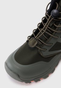 Botte de randonnée montante vert foncé avec semelle épaisse, maintenue par des lacets élastiques et un système de blocage à bascule, présentée sur un fond clair.