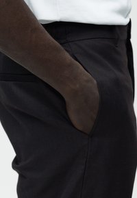 Χέρι με σκούρο χρώμα δέρματος που ξεκουράζεται μέσα στην εμπρόσθια τσέπη μαύρου παντελονιού, φορώντας άσπρο πουκάμισο μέσα στο παντελόνι.