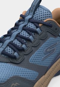 Κοντινή λήψη αθλητικού παπουτσιού με δίχτυ σε μπλε και ναυτικό χρώμα, κορδόνια με σχέδιο, επενδεδυμένο κολάρο και μπεζ σόλα με λογότυπο "S" στο πλάι.