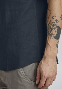 Cămașă din in de culoare albastru închis, cu o croială relaxată, despicătură pe lateral și un branding subtil, combinată cu pantaloni de un bej deschis, care expun o mână tatuată.