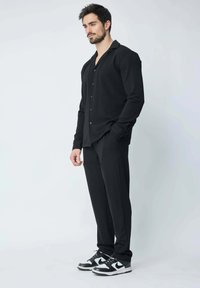Chemise noire à manches longues avec boutons, associée à un pantalon noir et des baskets noires avec des accents blancs. Texture lisse sur l'ensemble.
