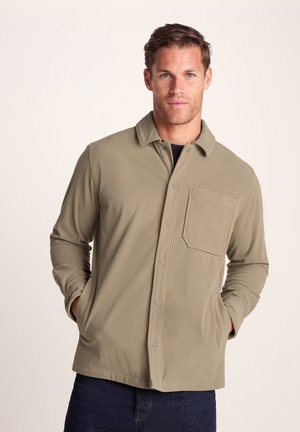 Man wearing a beige corduroy button-up jacket with front pocket, hands in pockets, standing against a plain light background.