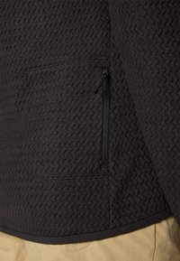 Primo piano di un giubbotto in pile nero con tessuto texturato, dotato di tasca verticale con zip, indossato sopra dei pantaloni beige.
