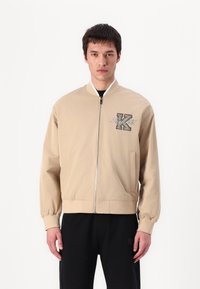Beige bomber jacket with a zip closure, ribbed cuffs, and hem. Features a black embroidered 'K' logo on the left chest. Smooth fabric texture.