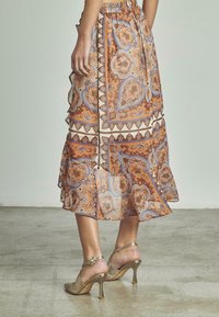 Jupe midi fluide à motifs en orange, bleu et crème, avec un ourlet à volants. Associée à des sandales à talons ouvertes et métalliques avec une bride.