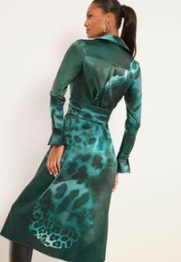 Trenci din satin verde cu imprimeu leopard. Are talie cu curea, mâneci lungi și guler, accentuând un design fluid.
