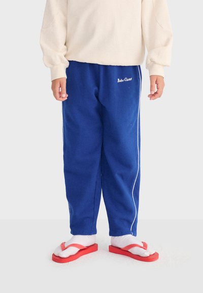 Niño con pantalones deportivos azules con franjas blancas en los costados, calcetines blancos, chanclas rojas y una camiseta de manga larga color crema, de pie sobre una superficie clara.