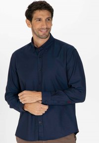 Chemise bleu marine à manches longues, avec un design ajusté et un détail de logo rouge discret sur le poignet gauche. Texture de tissu douce et lisse.