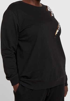 Zwarte trui met lange mouwen met gouden en zilveren pailletten bliksemschicht op de borst, gedragen met zwarte broek.