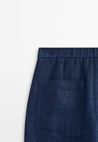 Section de pantalon en jean bleu foncé montrant une ceinture élastique et une grande poche arrière carrée sur fond blanc.